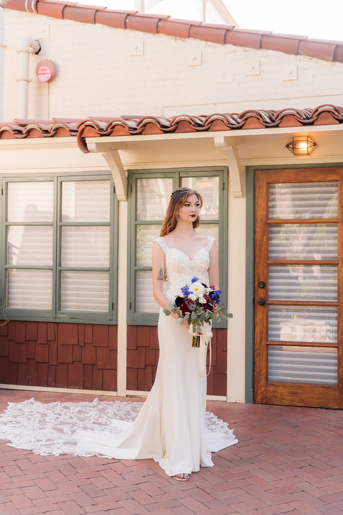 A bride in a sleek, modern wedding gown stands in front of a rustic cottage, holding a bouquet of vibrant flowers, showcasing the 2025 wedding photography trend of blending vintage charm with contemporary elegance.