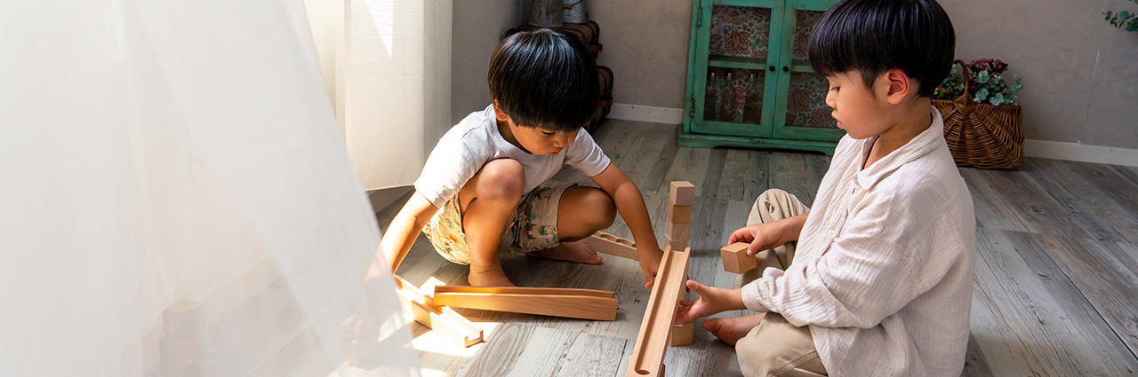 Two young children playing with wooden blocks on the floor in a naturally lit room, with soft sunlight streaming through a window covered by sheer curtains.