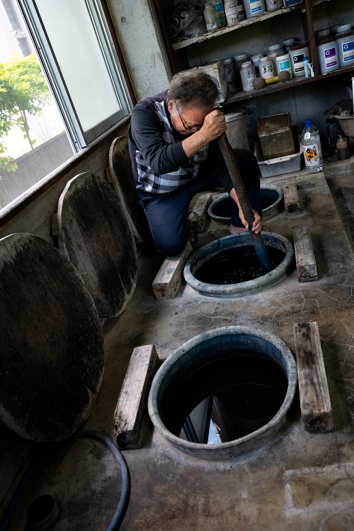 Um artesão agita cuidadosamente grandes cubas de tintura de índigo em uma oficina rústica, com luz natural entrando por uma janela próxima, iluminando o espaço de trabalho de madeira e concreto repleto de ferramentas e materiais.