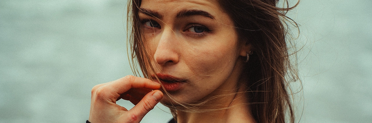 A close-up portrait of a woman with windblown hair, gazing into the camera with an intense expression. The image has been enhanced using color grading techniques to create a cinematic, moody atmosphere with soft contrast and cool tones.