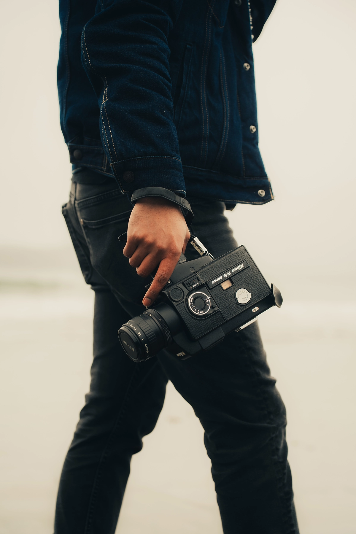 Gros plan d'une personne portant une veste en jean et un jean noir, tenant un appareil photo vintage à ses côtés. L'image a été améliorée à l'aide de techniques d'étalonnage des couleurs pour obtenir une esthétique chaleureuse et cinématographique avec un contraste doux.