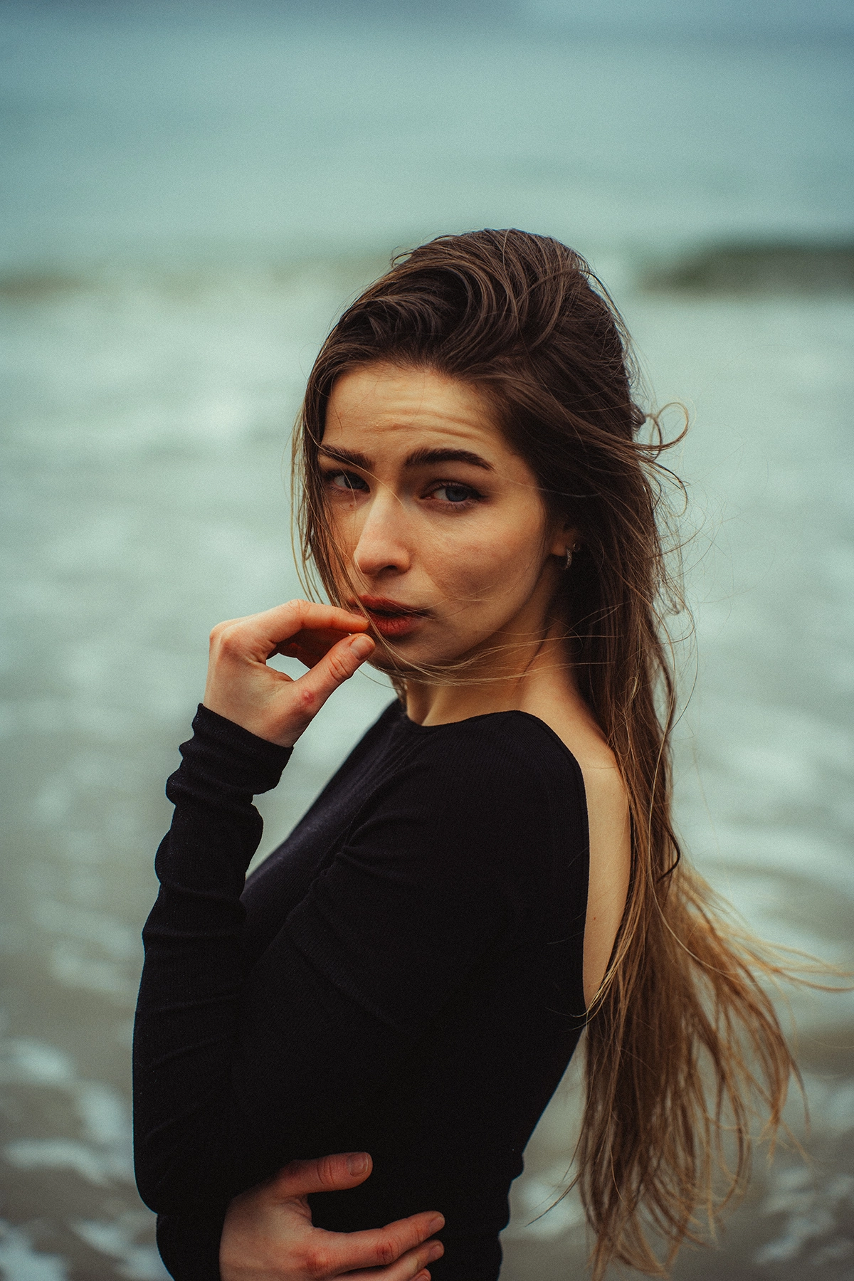 Portrait d'une femme debout au bord de l'océan, les cheveux balayés par le vent, fixant intensément l'appareil photo. L'image a été améliorée à l'aide de techniques d'étalonnage des couleurs pour créer une atmosphère cinématographique et morose avec des tons profonds et un contraste riche.