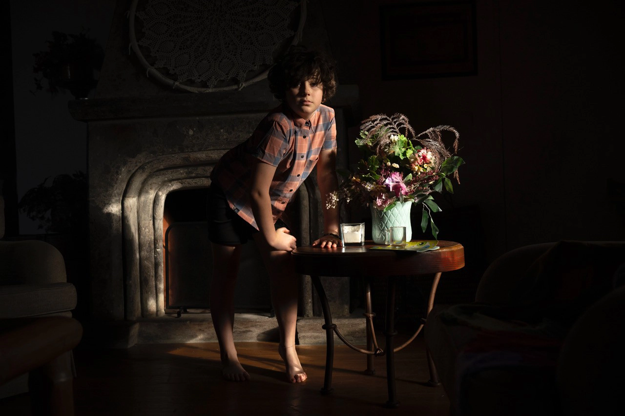 A moody indoor portrait of a child lit by early morning light, standing near a table with a floral arrangement—an example of photographing portraits in early morning light using natural window illumination.
