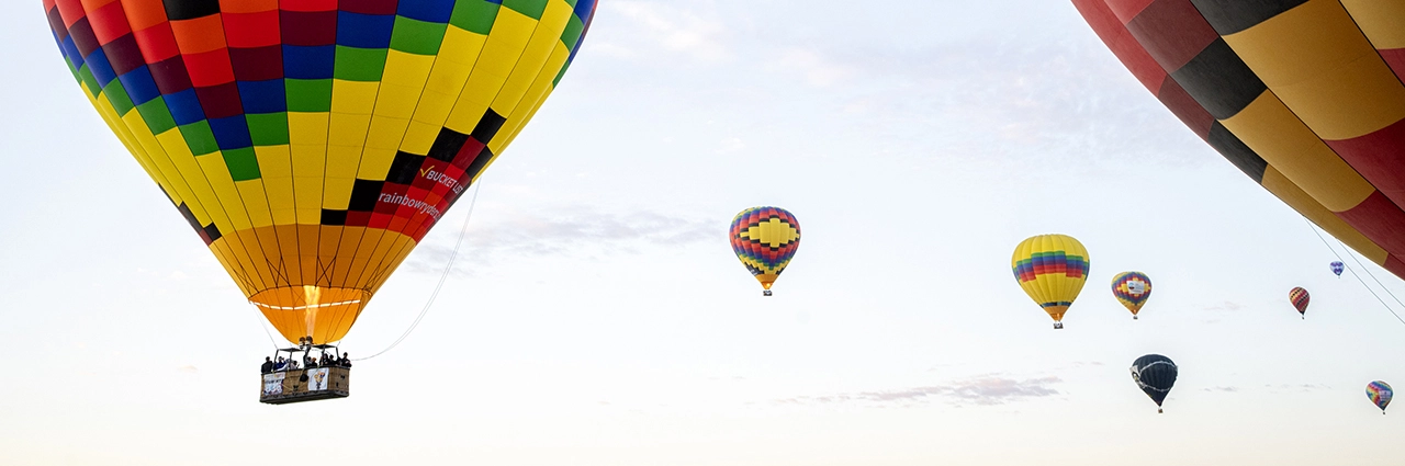 Multiple colorful hot air balloons floating in a clear sky during a balloon festival, with a large, vibrant balloon carrying passengers in the foreground.