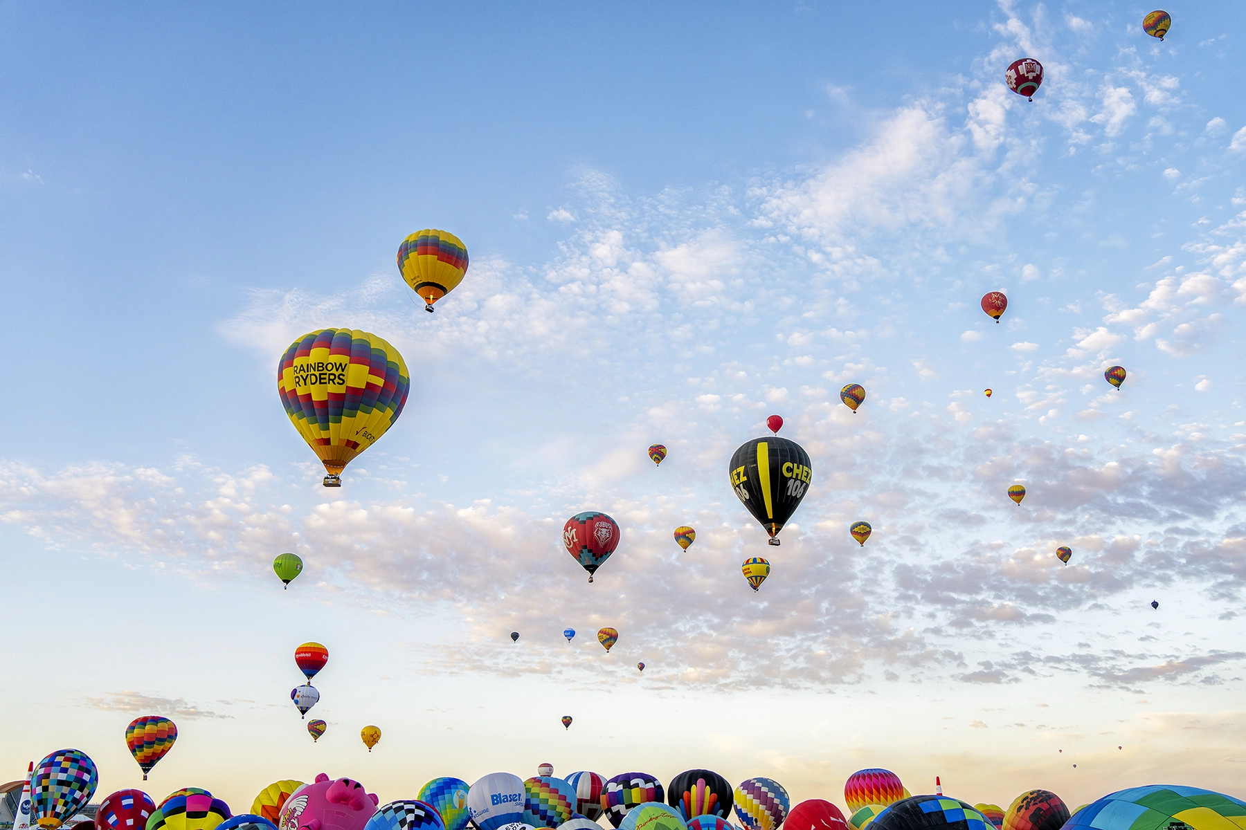 Hot air balloons of various colors and designs ascending into a partly cloudy sky during a balloon festival, with the 'Rainbow Ryders' balloon prominently featured
