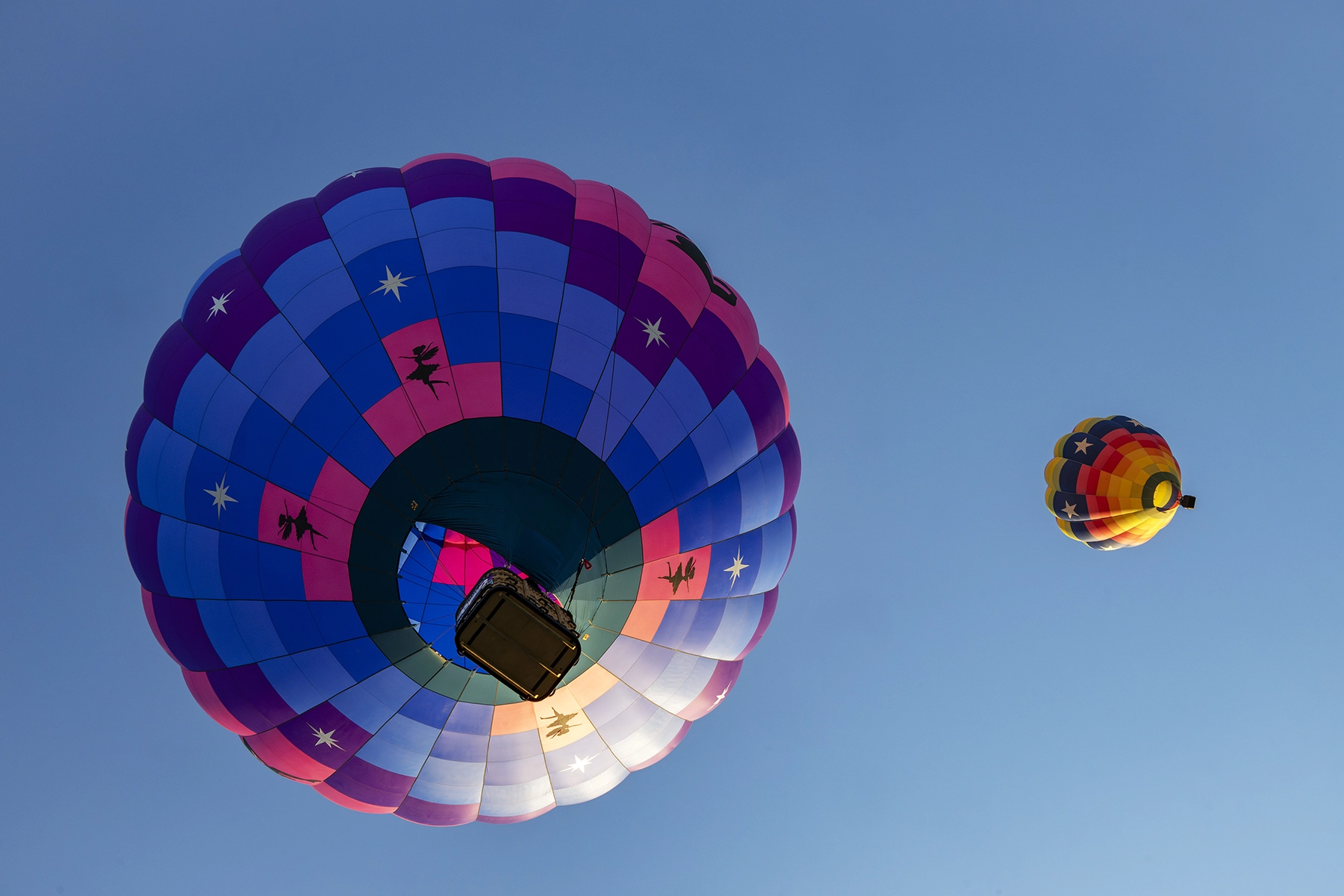 Two colorful hot air balloons viewed from below against a clear blue sky, with one featuring star and silhouette designs and the other showcasing a rainbow pattern.