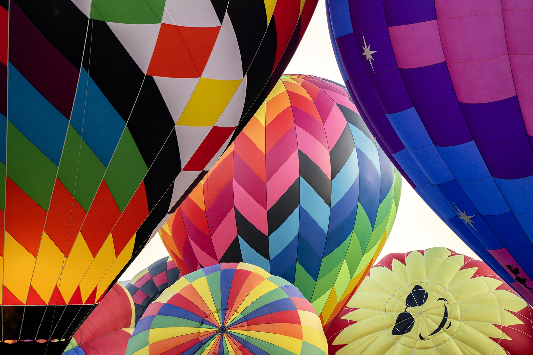 Close-up of colorful hot air balloons with various patterns, including stripes, chevrons, and a smiling sun design, clustered together at a balloon festival.