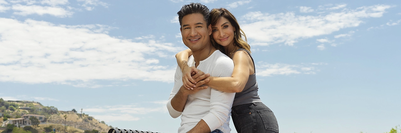 Un couple souriant s'embrasse en plein air sous un ciel bleu lumineux, avec une colline pittoresque en arrière-plan. Des réflecteurs ont été utilisés pour renforcer la lumière naturelle, créant ainsi un portrait chaleureux et équilibré.