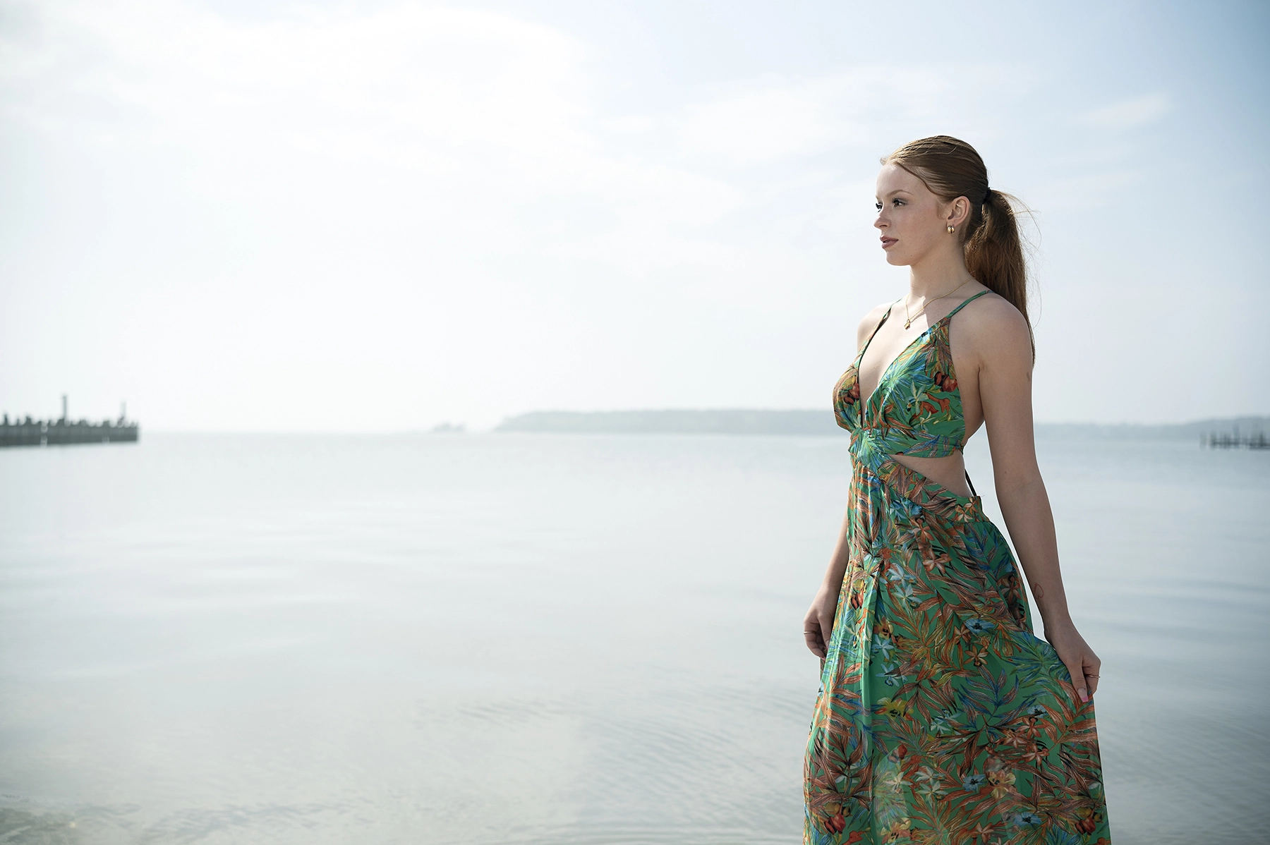 Une femme vêtue d'une robe à l'imprimé tropical se tient au bord de l'océan et regarde au loin. Le portrait à contre-jour a été mis en valeur par des réflecteurs pour équilibrer la lumière naturelle et créer une composition douce et bien éclairée.