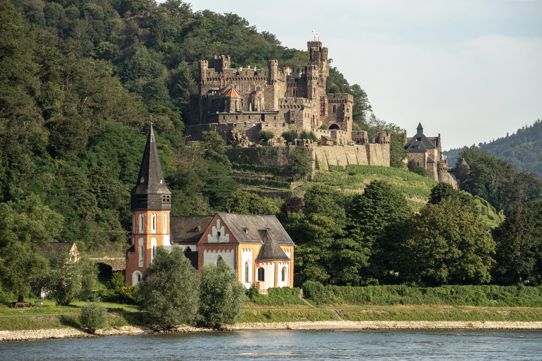Rheinstein Castle and a riverside church surrounded by lush greenery along the Rhine River in Germany.