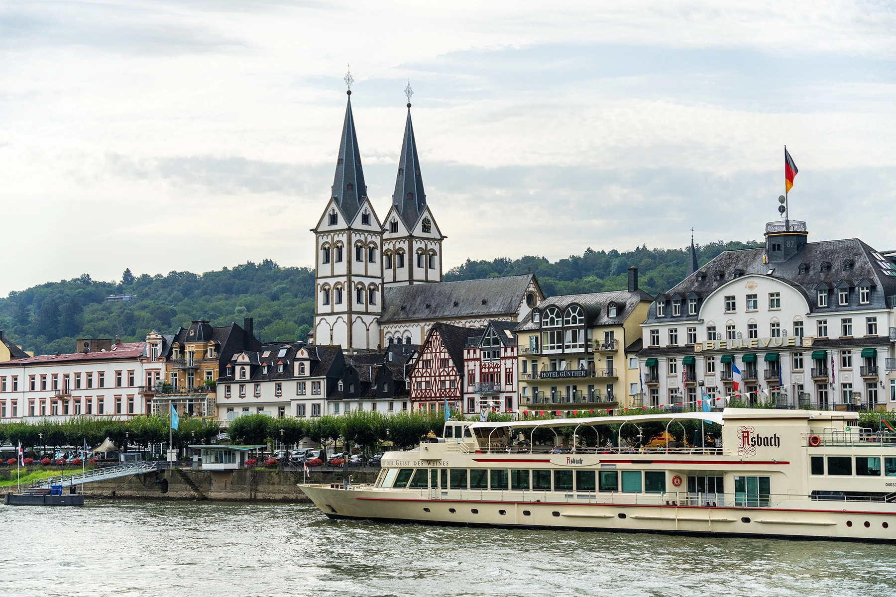 A river cruise boat passing by historic buildings and a church with twin spires in Boppard, Germany. Images like this a plentiful when engaging in Rhine River cruise travel photography.