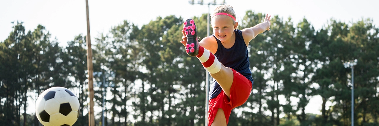 Panoramic action shot of a young girl kicking a soccer ball with intensity, framed to emphasize motion and energy in a bright outdoor sports setting.