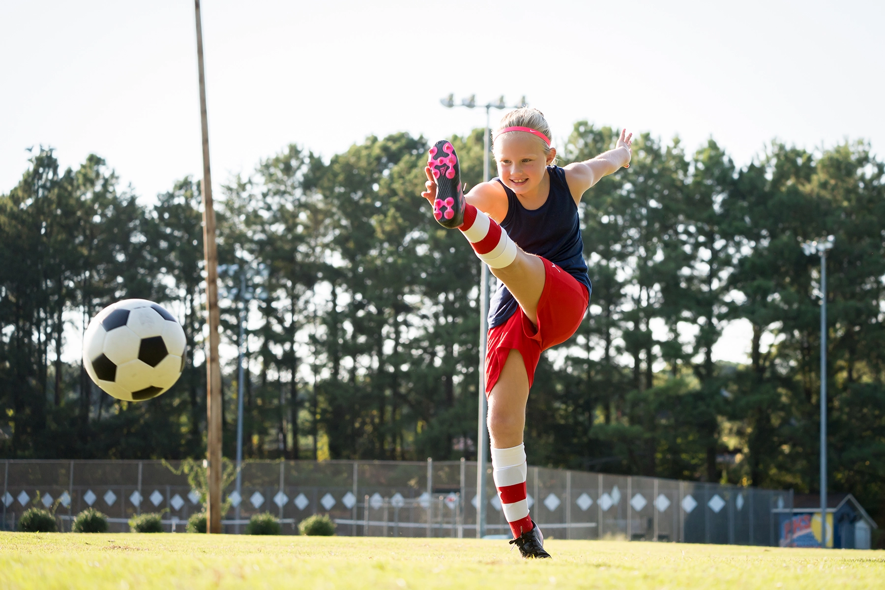 Young girl kicking a soccer ball mid-action on a sunny field, captured with a fast shutter speed and mid-range F-number to freeze motion and maintain depth of field.