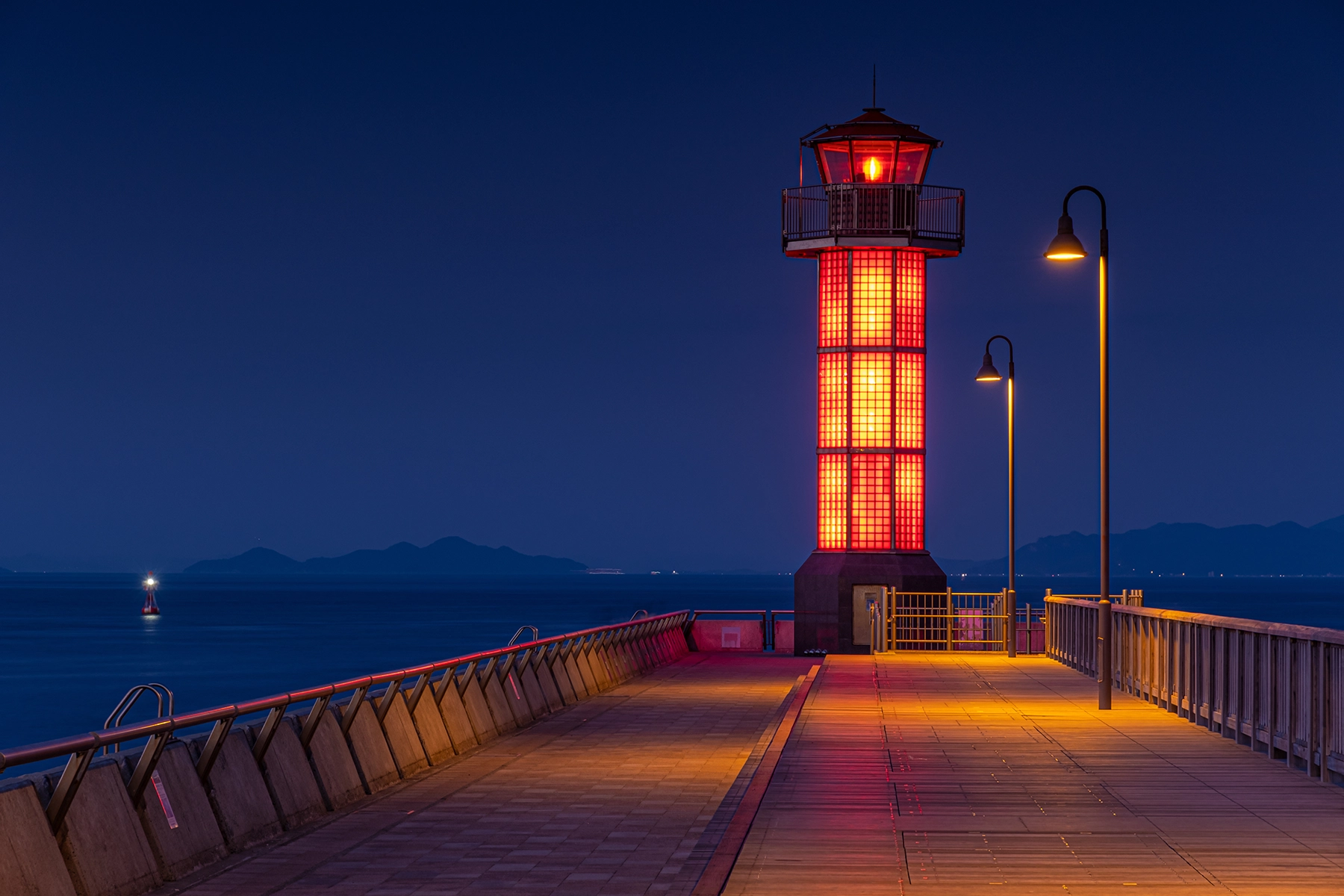 Night view of a red-lit lighthouse on a pier with distant mountains, captured with a long exposure and high F-number to ensure clarity and depth in low-light conditions.