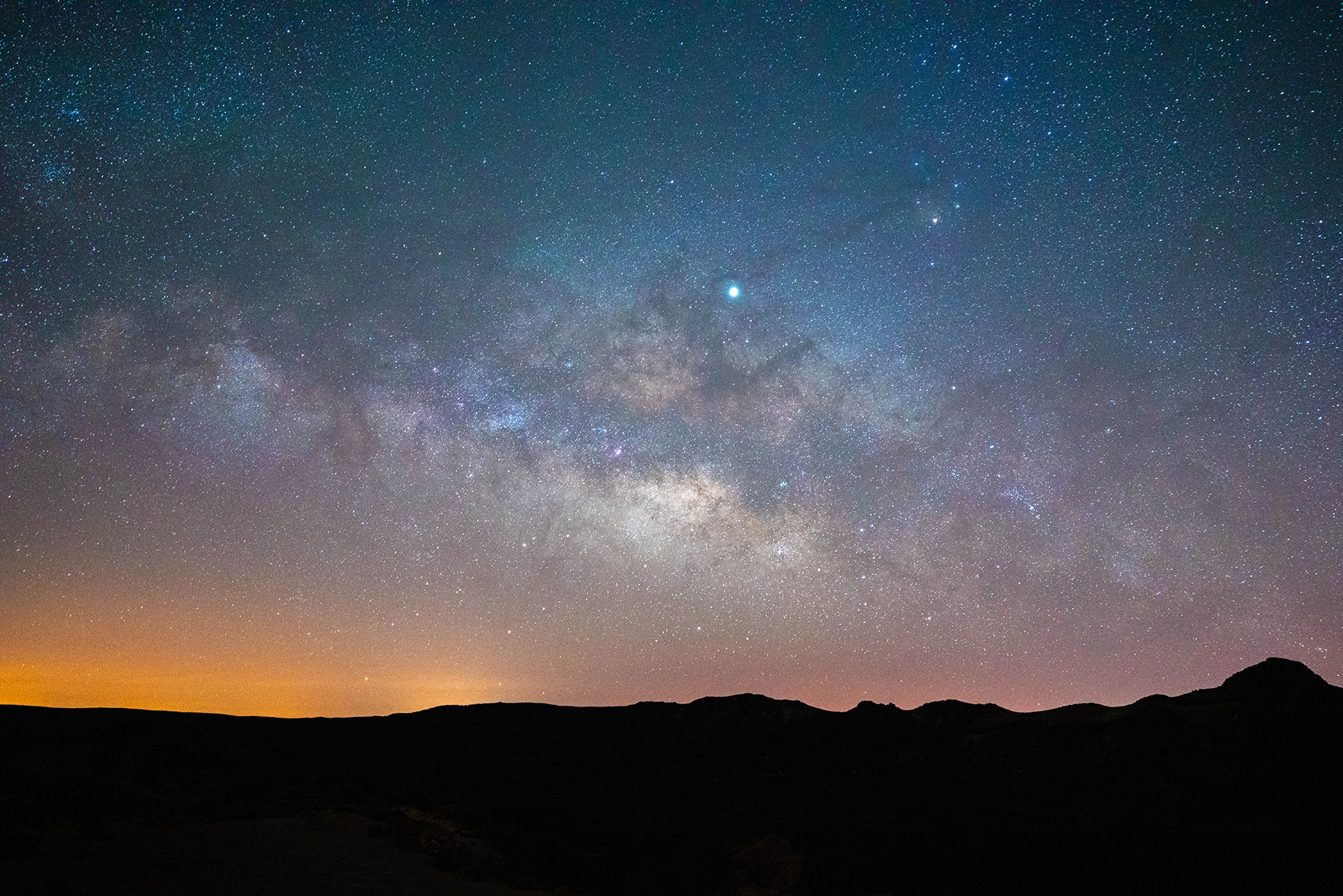 Panoramic night sky with a glowing Milky Way over silhouetted mountain ridges, captured with a wide-angle lens and wide aperture to gather maximum starlight.