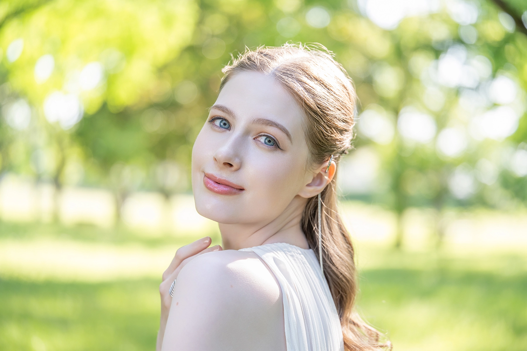 Portrait of a woman in soft sunlight with a serene expression, captured outdoors with lush green trees in the background blurred into soft circular bokeh, highlighting the use of natural background bokeh in portrait photography.