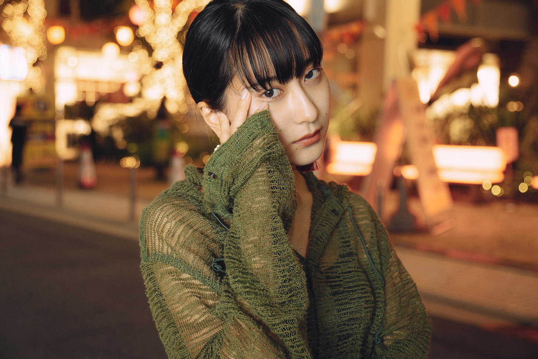 Woman in a green textured outfit posing on a city street at night, with blurred background lights forming soft bokeh, creating a dramatic and atmospheric urban portrait.