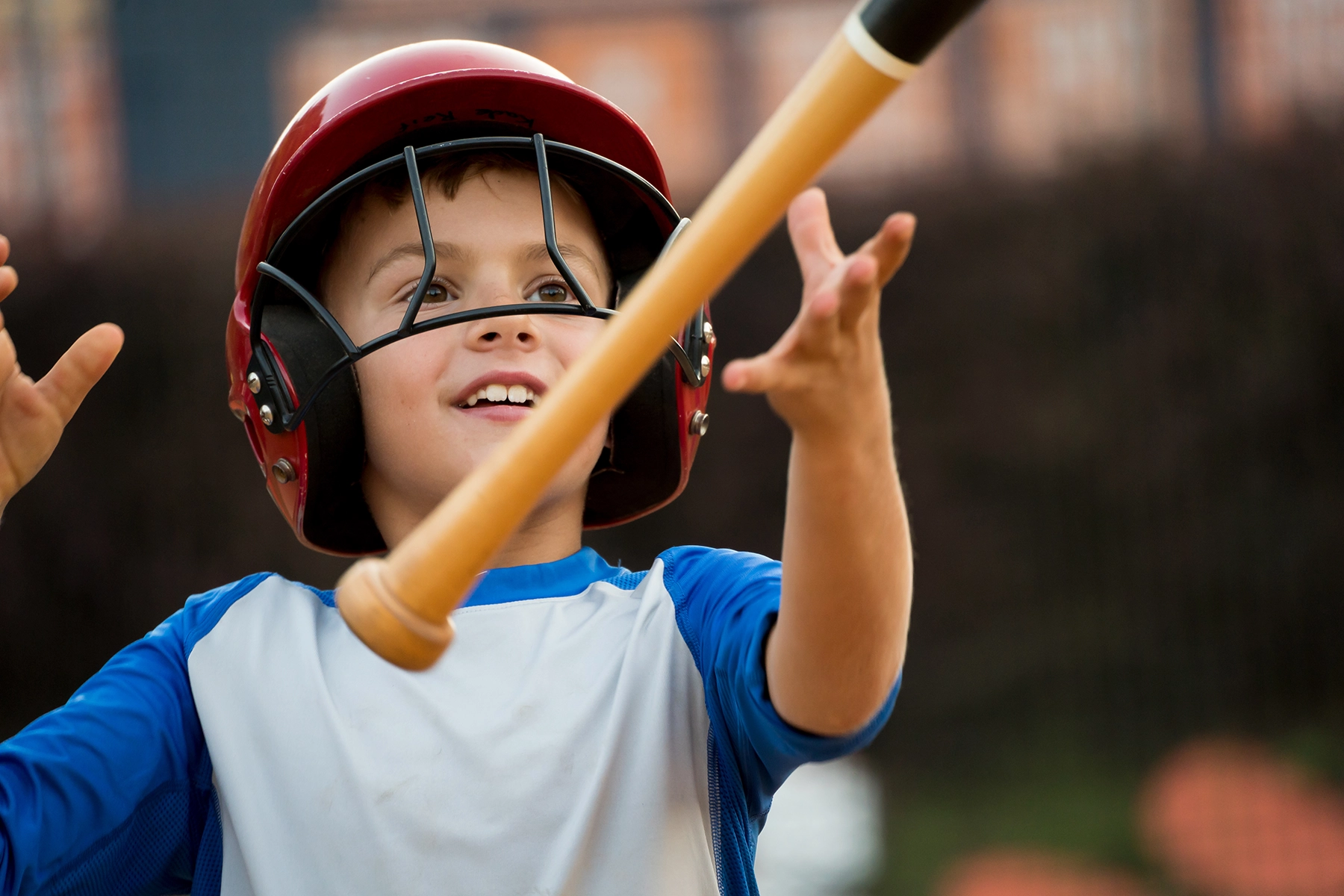 Smiling young baseball player in a red helmet tossing a bat in the air, captured with a telephoto lens to highlight expression and motion against a softly blurred background.