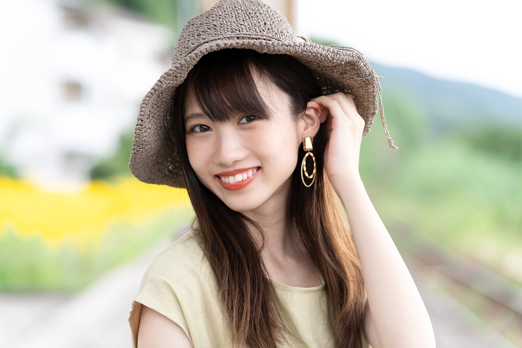 Smiling woman in a sunhat and gold earrings posing outdoors, captured with a medium telephoto focal length for soft background bokeh and subject isolation.