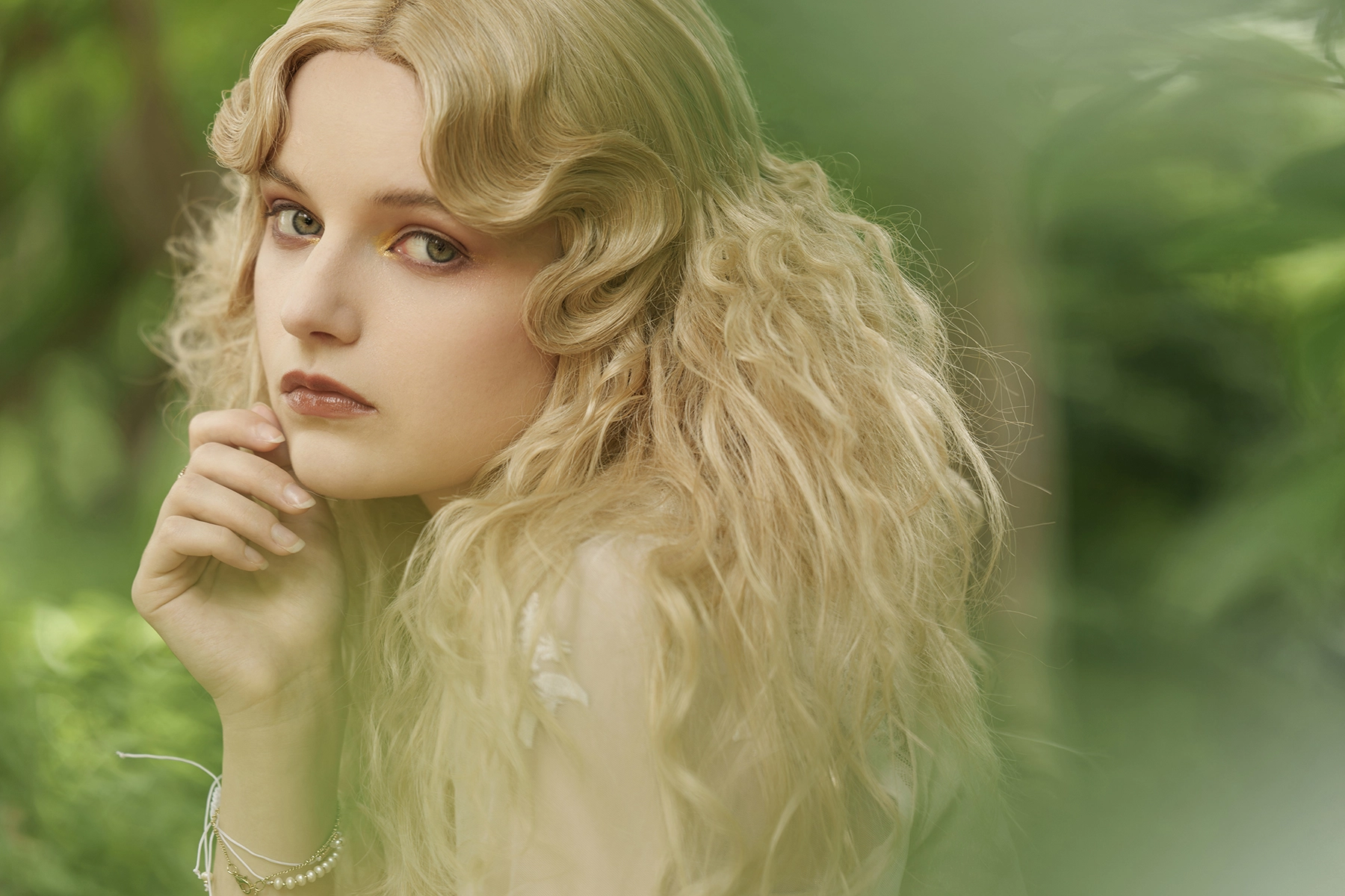 Close-up portrait of a woman with wavy blonde hair in soft natural light, photographed through foliage using a telephoto lens for shallow depth of field and smooth background bokeh.