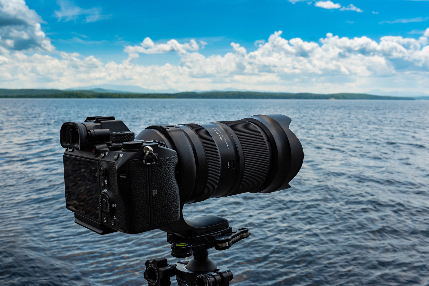 Telephoto lens mounted on a mirrorless camera and tripod, set up by a lake under a bright sky, ready to capture distant landscapes.