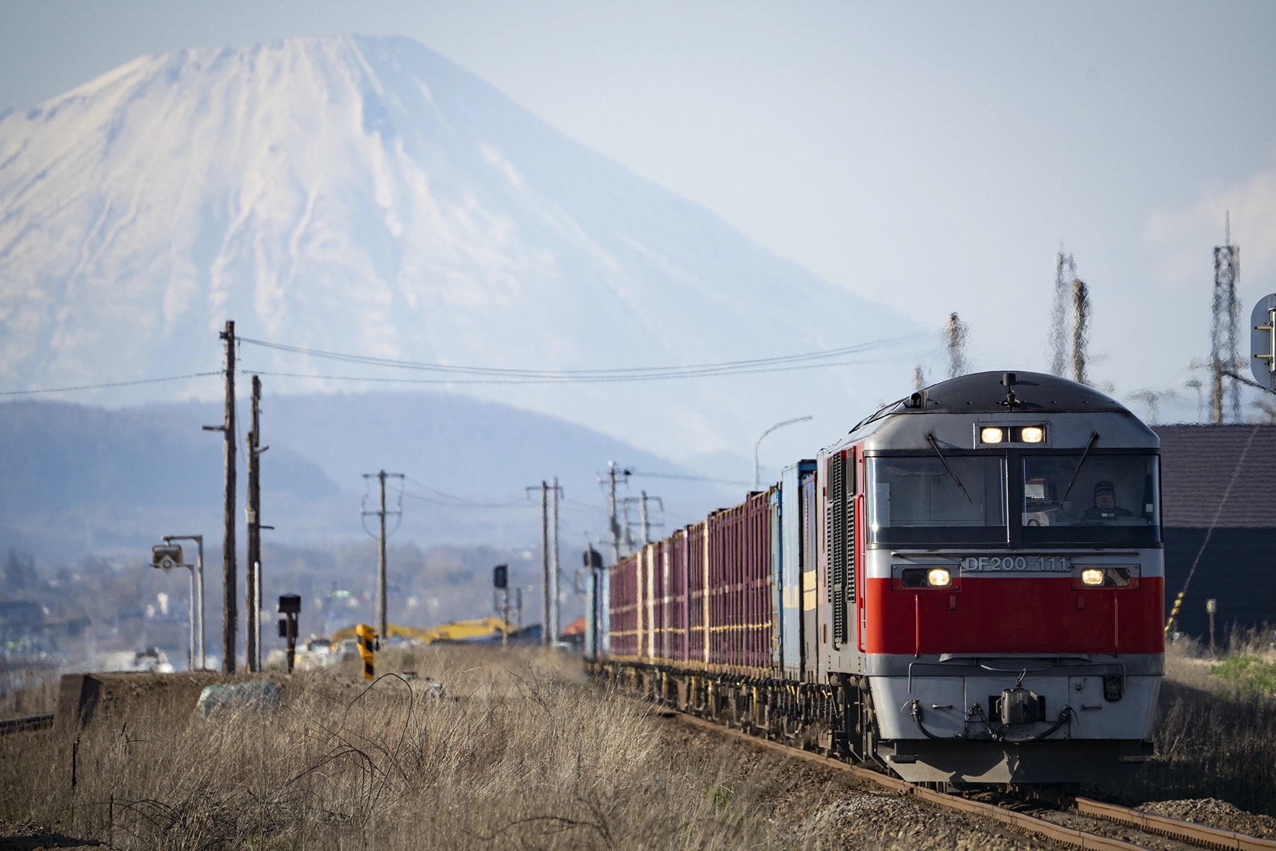 A freight train approaches on a railway track with a snow-covered mountain dramatically compressed in the background, demonstrating the magnified perspective effect of a telephoto lens