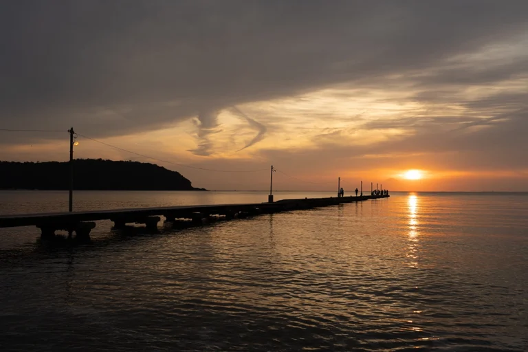 A dock jutting out into the water with the sun setting through the clouds