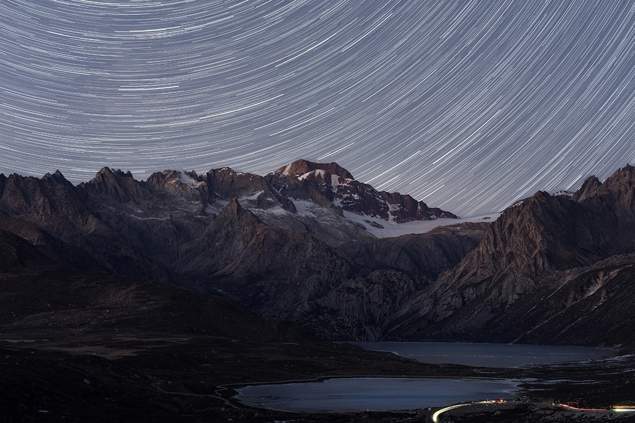 Star trails swirl above rugged mountain peaks and glacial lakes, captured through astrophotography with a macro lens—revealing fine sky details over Sister Lake.