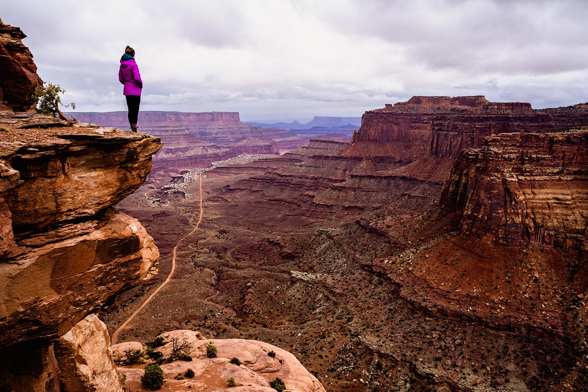 Una mujer vestida con una chaqueta púrpura se encuentra al borde de un acantilado rocoso, con vistas al vasto paisaje del Gran Cañón bajo un cielo nublado-capturado utilizando un objetivo de cámara para fotografía de montaña para resaltar el terreno dramático y la profundidad distante.