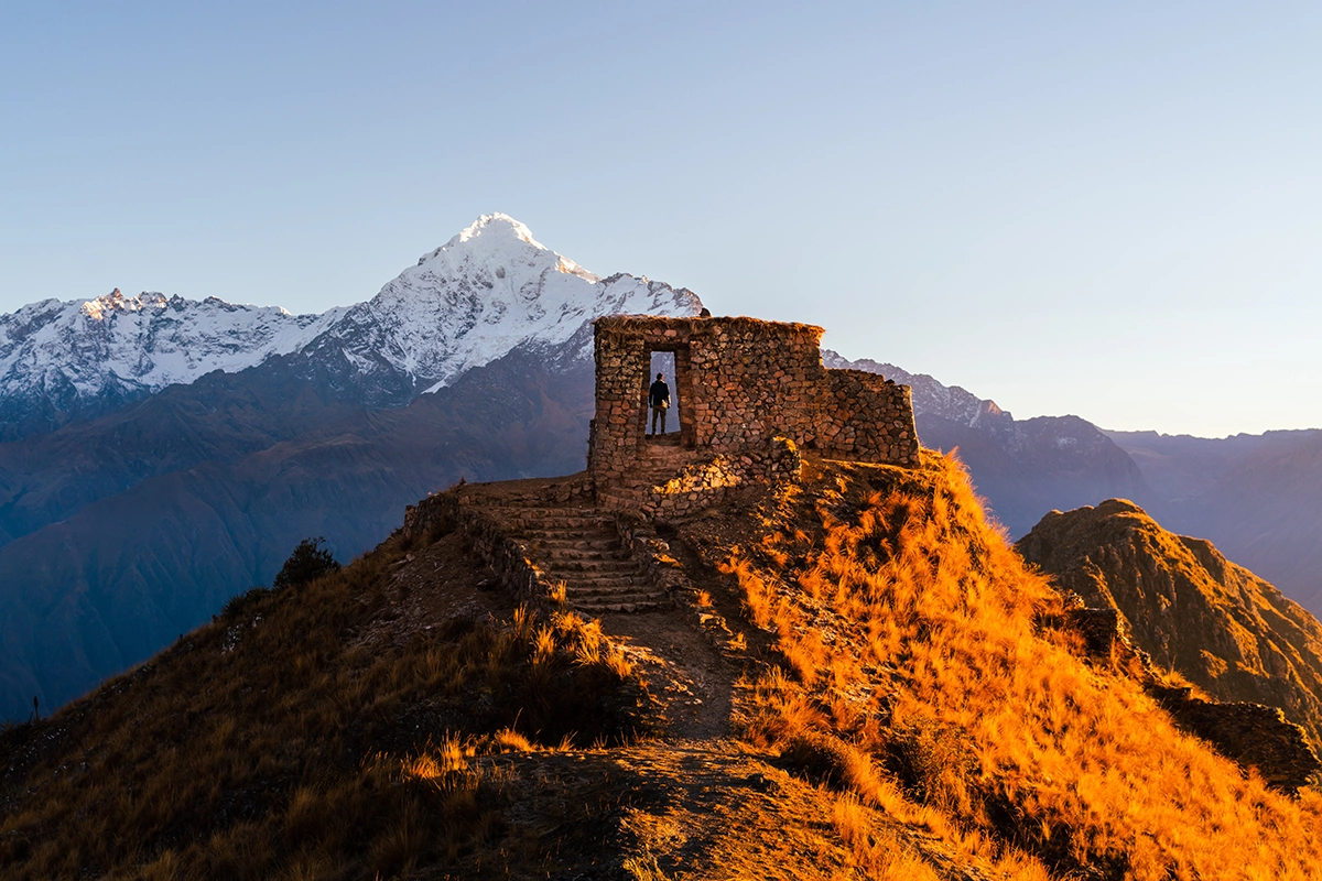 Un viajero solitario se encuentra en el interior de unas ruinas de piedra al amanecer, enmarcado por la luz dorada de los Alpes y una montaña nevada como telón de fondo, captada con un objetivo de fotografía de montaña para resaltar la textura, la escala y la elevación.