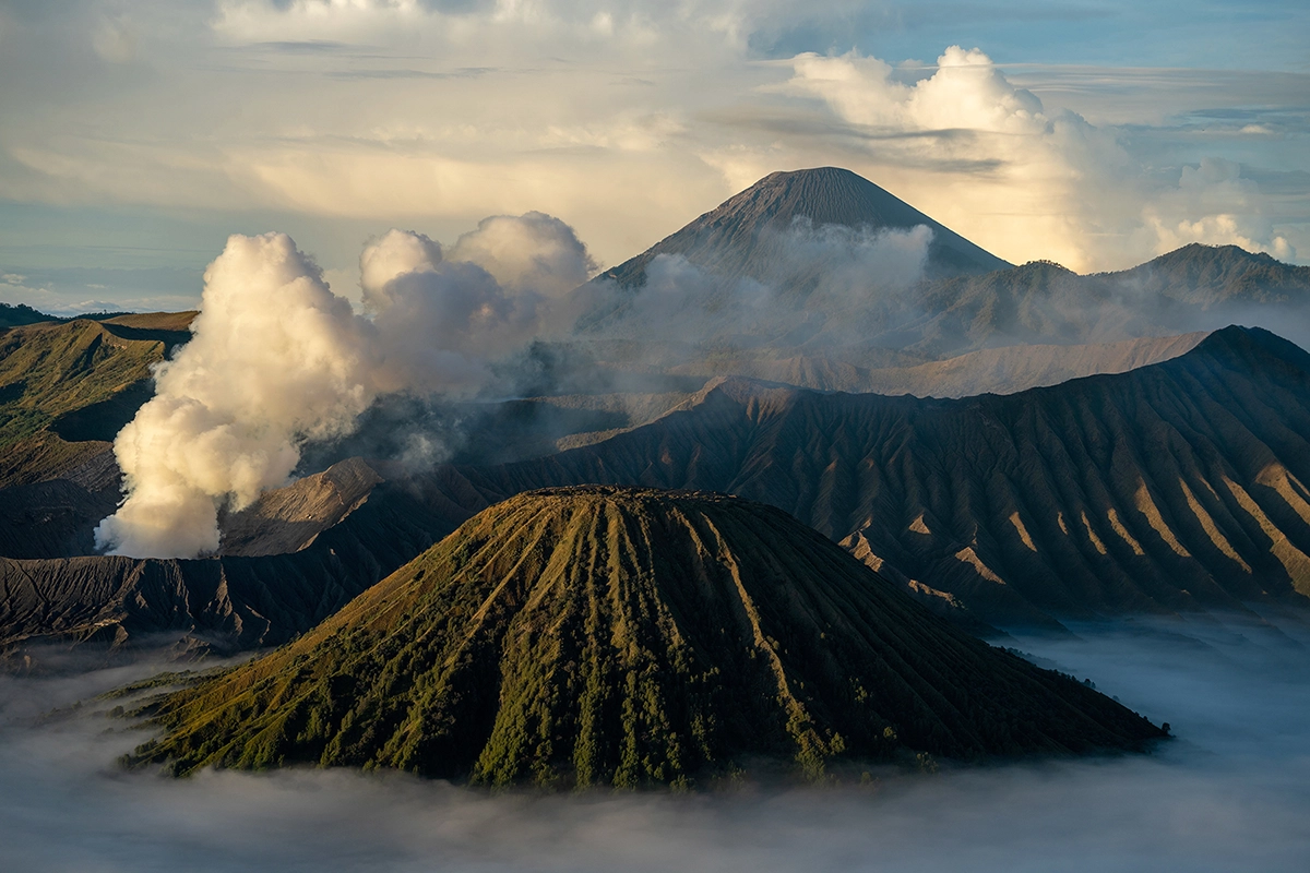 Un paisaje volcánico en capas con humo saliendo del monte Bromo al amanecer, enmarcado por la niebla y una luz suave, capturado con un objetivo para fotografía de montaña que revela texturas espectaculares, profundidad y detalles atmosféricos.