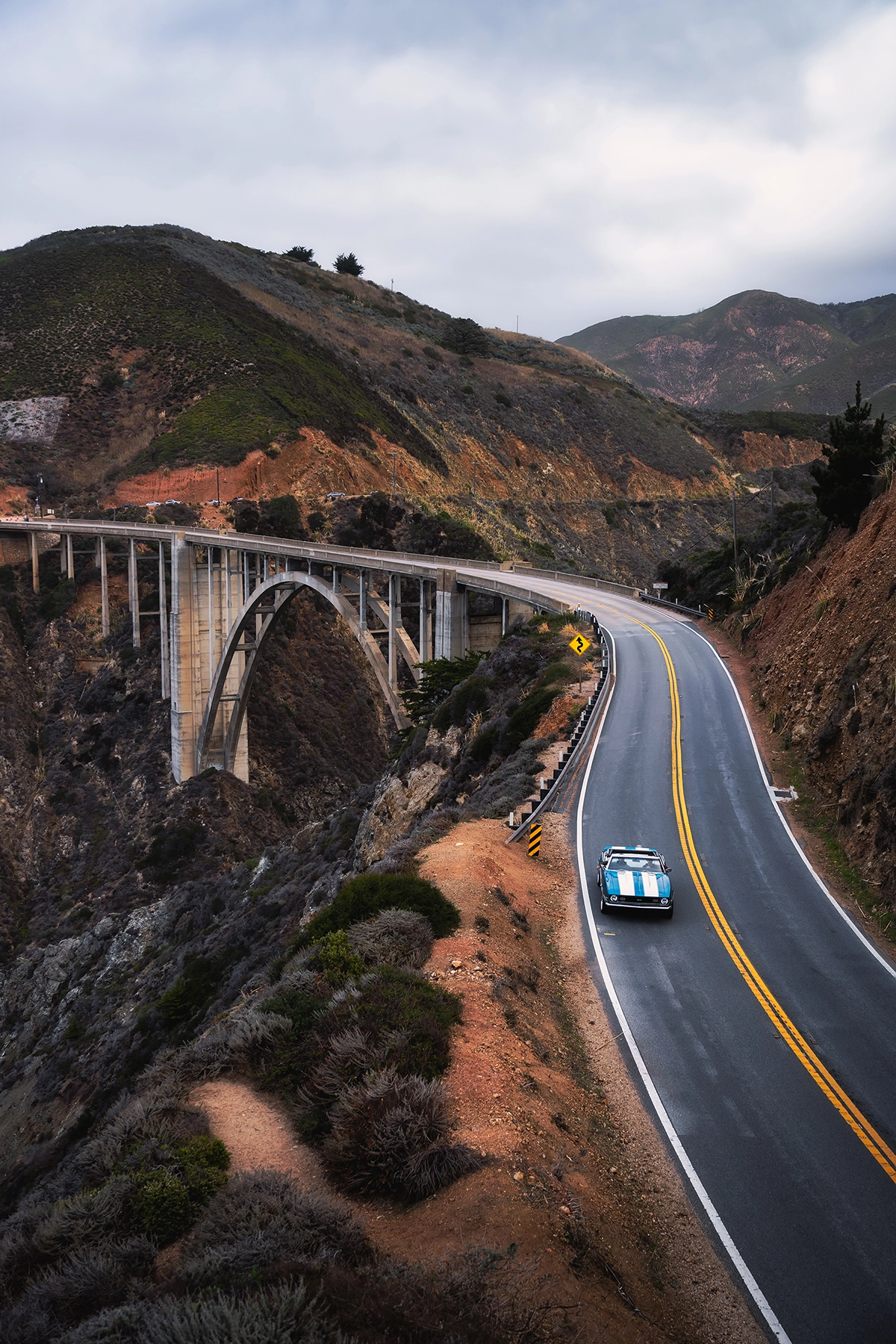 Clásico Camaro azul y blanco circulando por la autopista 1 cerca del puente de Bixby Creek en Big Sur, California, con acantilados costeros y escarpadas colinas de fondo.