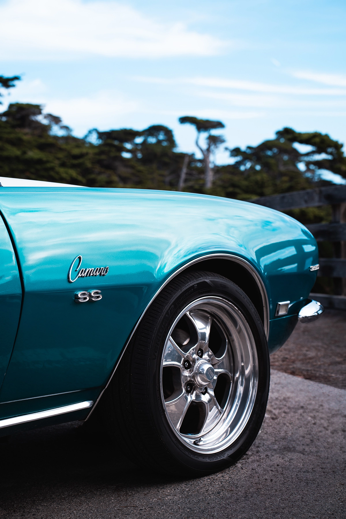 Primer plano de un Chevrolet Camaro SS turquesa aparcado en la costa de California, con detalles cromados en las ruedas y árboles costeros de fondo.