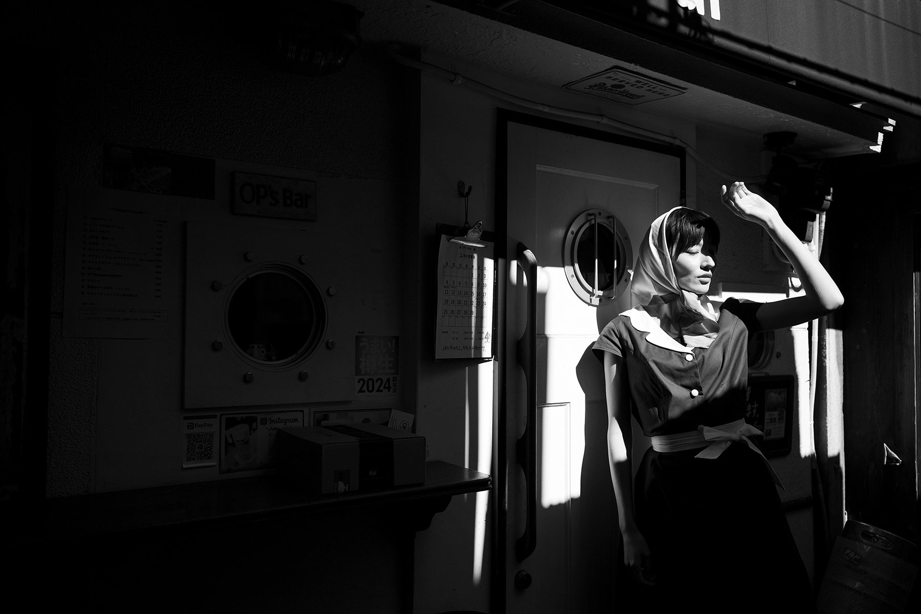 A woman wearing a vintage-style headscarf and dress stands outside a bar, shielding her eyes from the sunlight, with dramatic shadows cast on the wall in this black-and-white photograph.