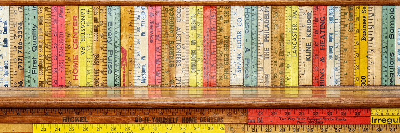 A creative bookshelf display made from colorful vintage rulers, showcasing a variety of advertising imprints from hardware and service businesses—an example of abstract art photography and unconventional subjects found at Luna Parc.