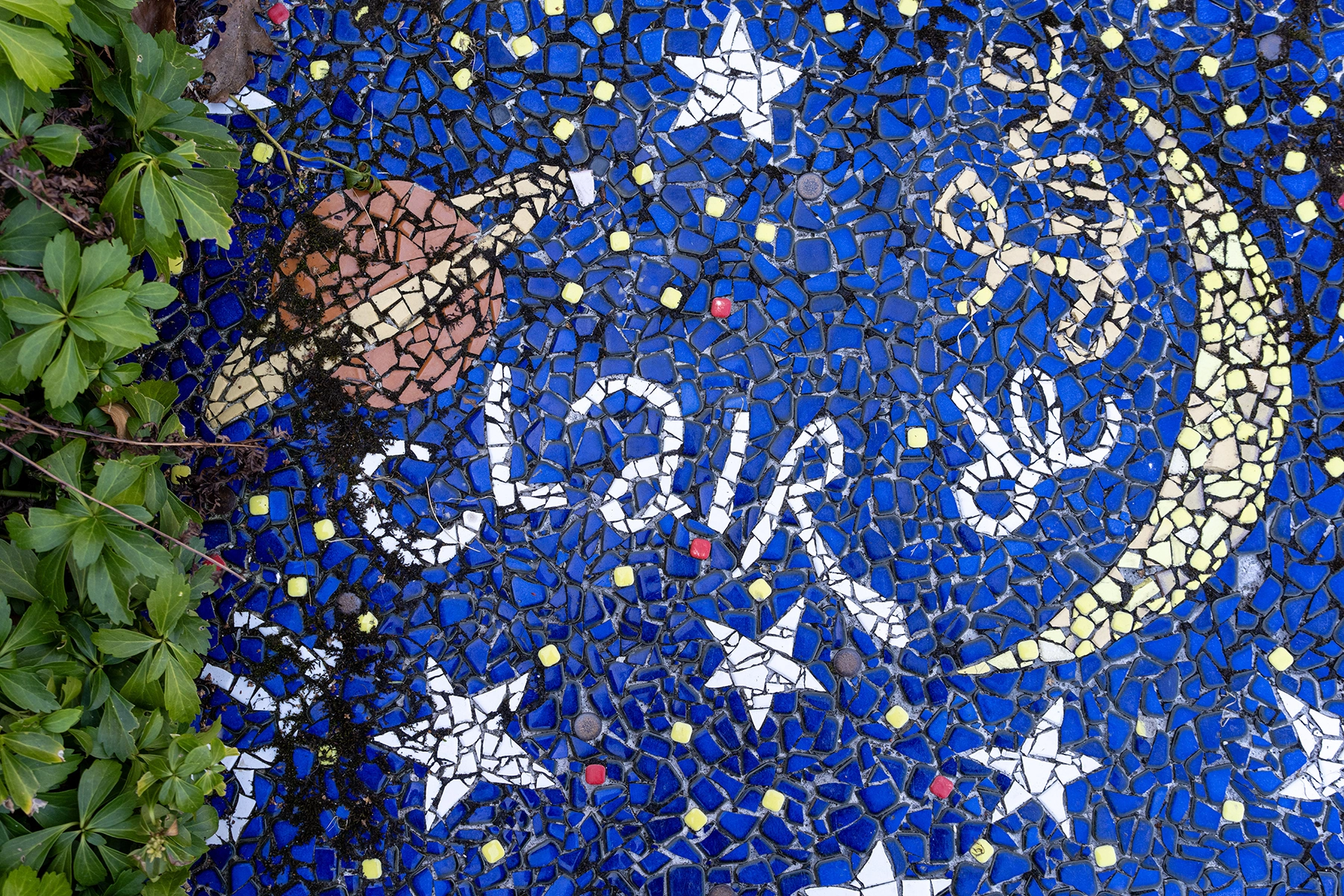 Colorful mosaic artwork at Luna Parc featuring a crescent moon, stars, Saturn, and abstract text on a deep blue background, partially bordered by green foliage—an example of photographing artistic surfaces and vibrant color patterns.