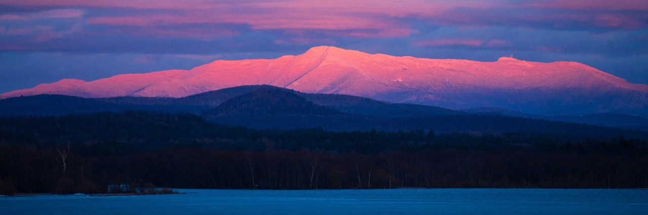 Un impresionante paisaje invernal con una montaña nevada bañada en suaves tonos rosas y morados por el sol poniente, con un lago helado y oscuras colinas boscosas en primer plano.