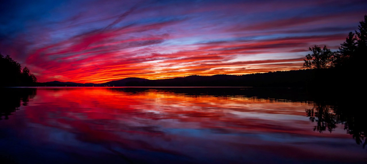 Una impresionante puesta de sol sobre un lago en calma, con vibrantes tonos rojos, naranjas y morados surcando el cielo y reflejándose maravillosamente en la superficie del agua.