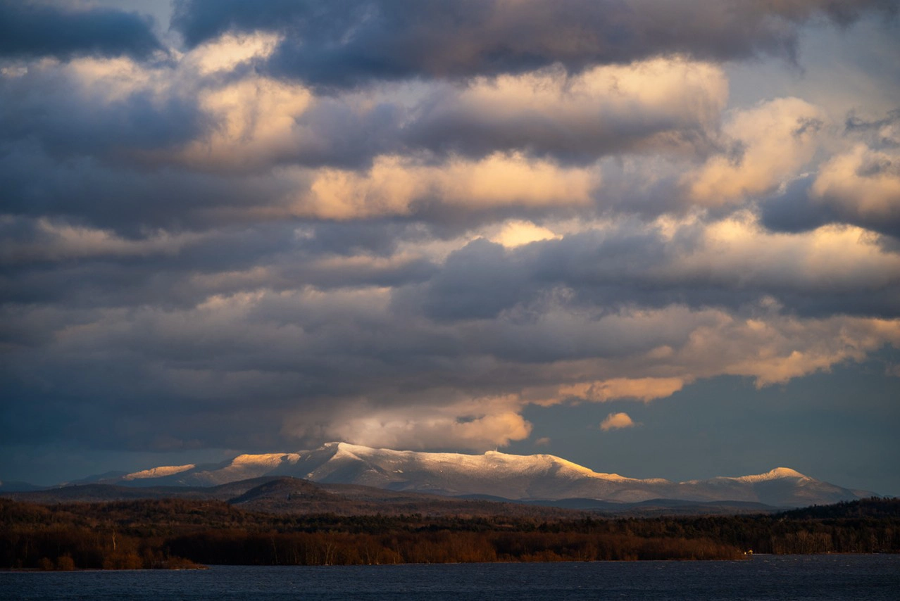 Un dramático paisaje de montañas nevadas iluminadas por la luz dorada del sol, parcialmente envueltas en densas nubes, con un lago oscuro y un bosque en primer plano.