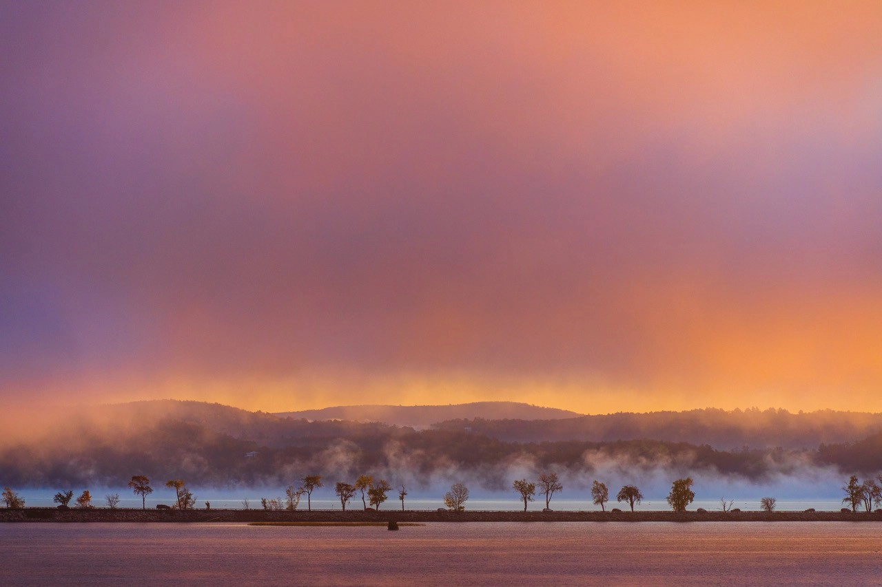 Un lago tranquilo al amanecer, con la bruma elevándose sobre el agua y un cielo colorido reflejándose en el paisaje.