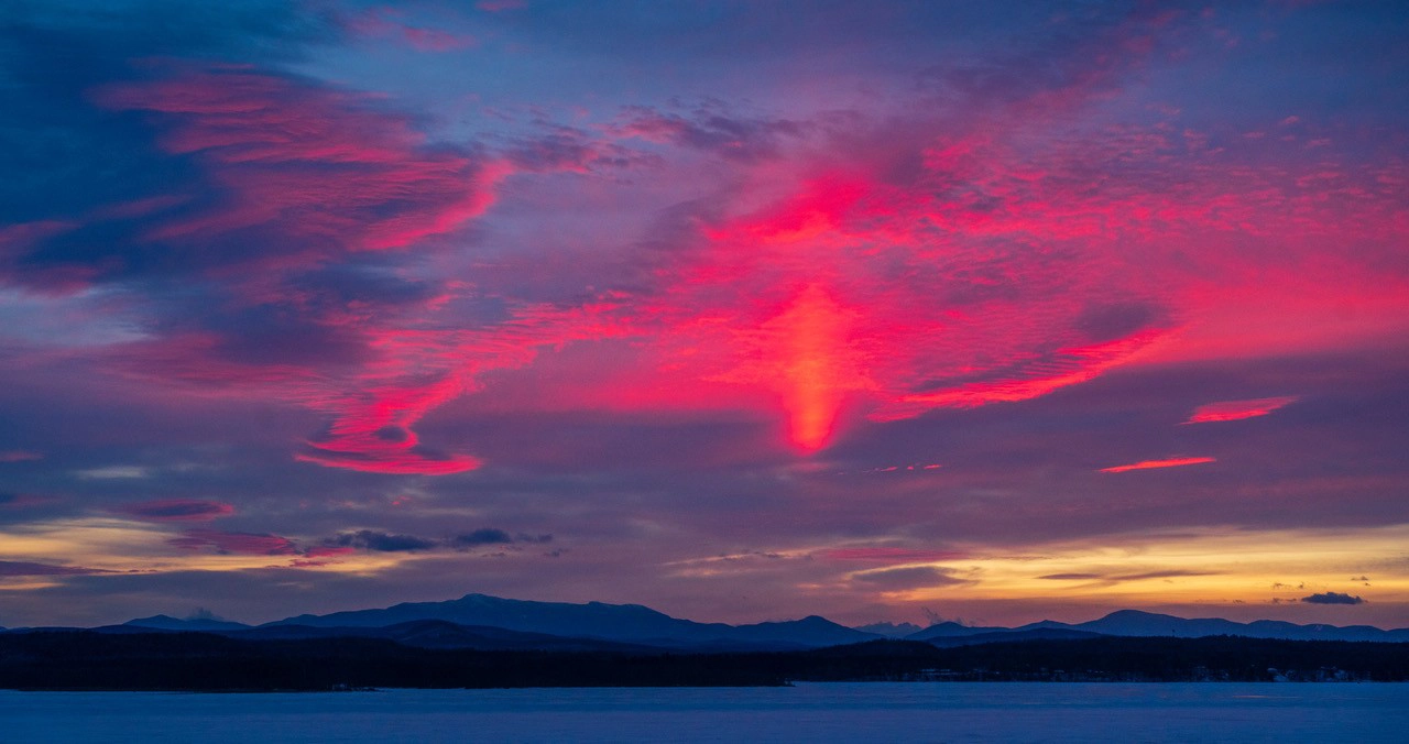 Una vibrante puesta de sol pinta el cielo en tonos rosas y morados sobre un paisaje montañoso y un lago helado.