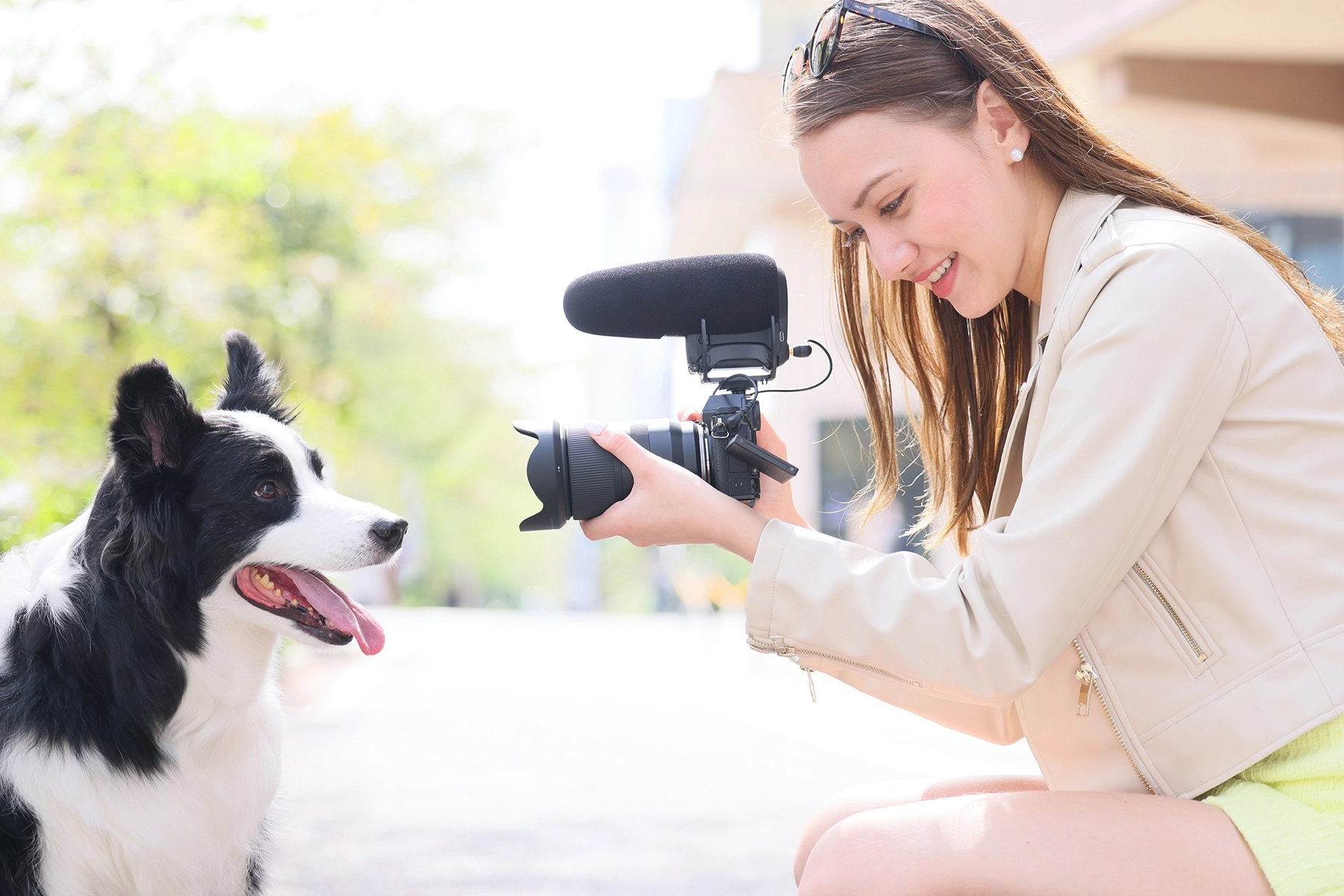 Una mujer sonriente filma a un Border Collie feliz al aire libre utilizando una cámara sin espejo con micrófono, mostrando una grabación de vídeo casual con luz natural.