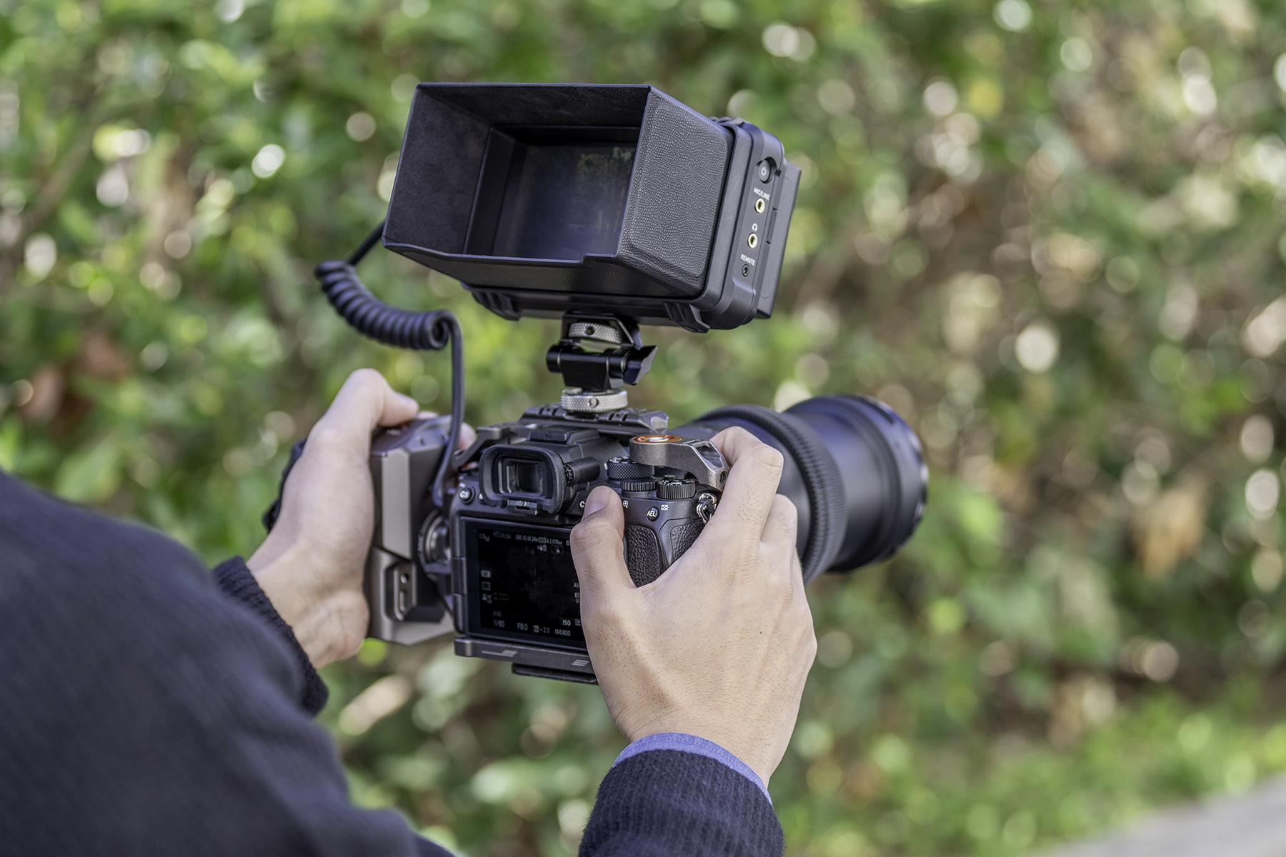 Fotógrafo usando uma câmera sem espelho com um monitor de campo externo e lente de zoom telefoto para gravação de vídeo ao ar livre.