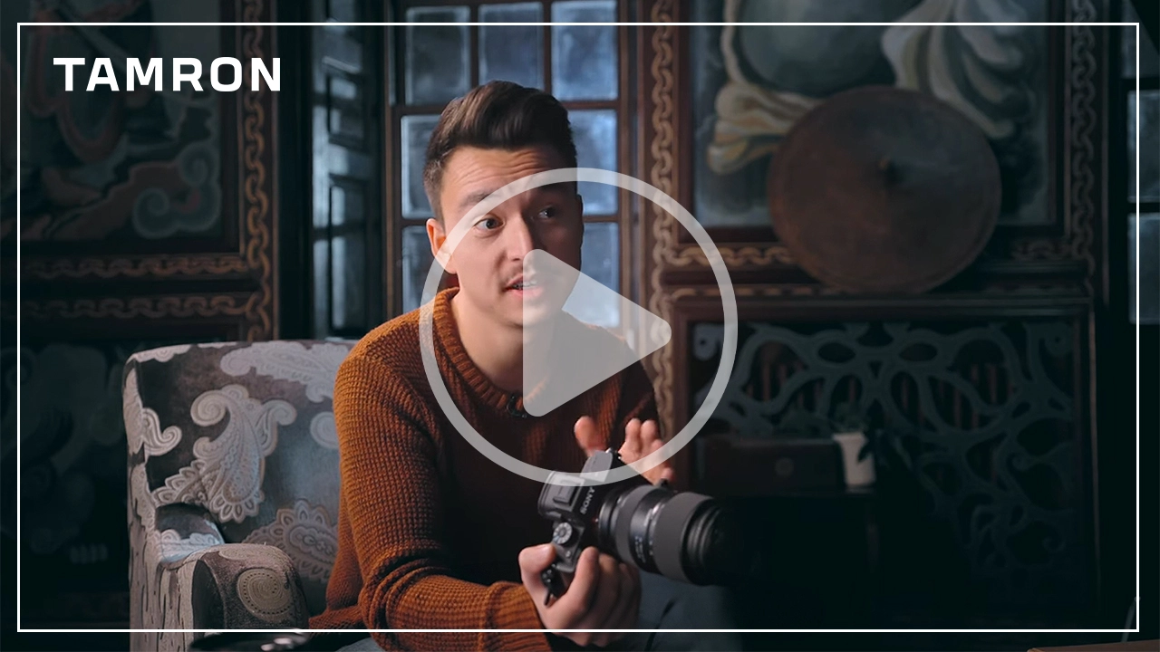 YouTuber assis à l'intérieur, tenant un appareil photo Sony équipé d'un objectif Tamron, discutant des caractéristiques du TAMRON Lens Utility Mobile, avec la marque Tamron et un bouton de lecture en surimpression.