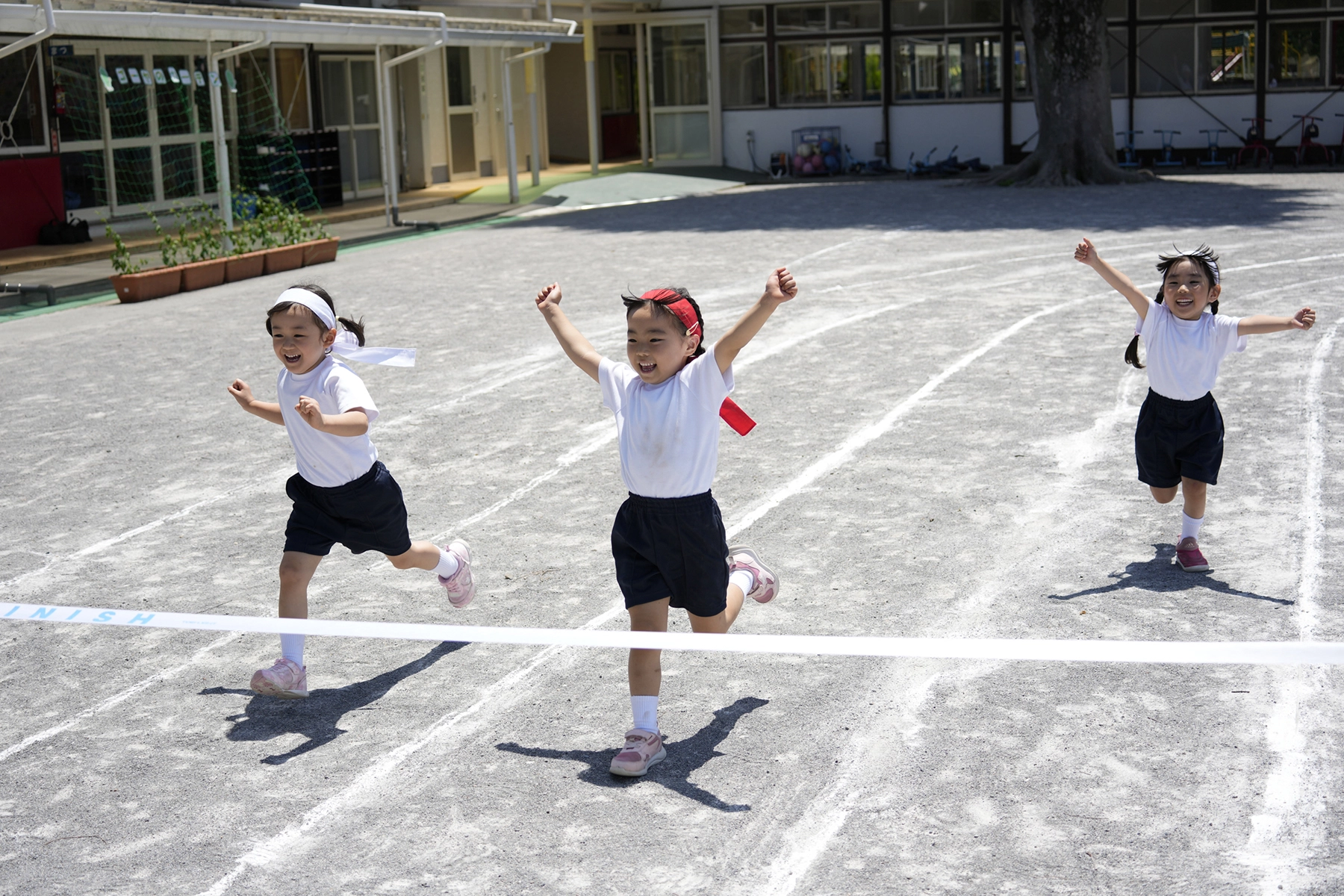 Un plan d'action montrant trois jeunes enfants qui courent joyeusement vers la ligne d'arrivée lors d'une journée sportive à l'école, les bras levés en signe d'excitation, sous un soleil radieux, sur une piste de gravier.