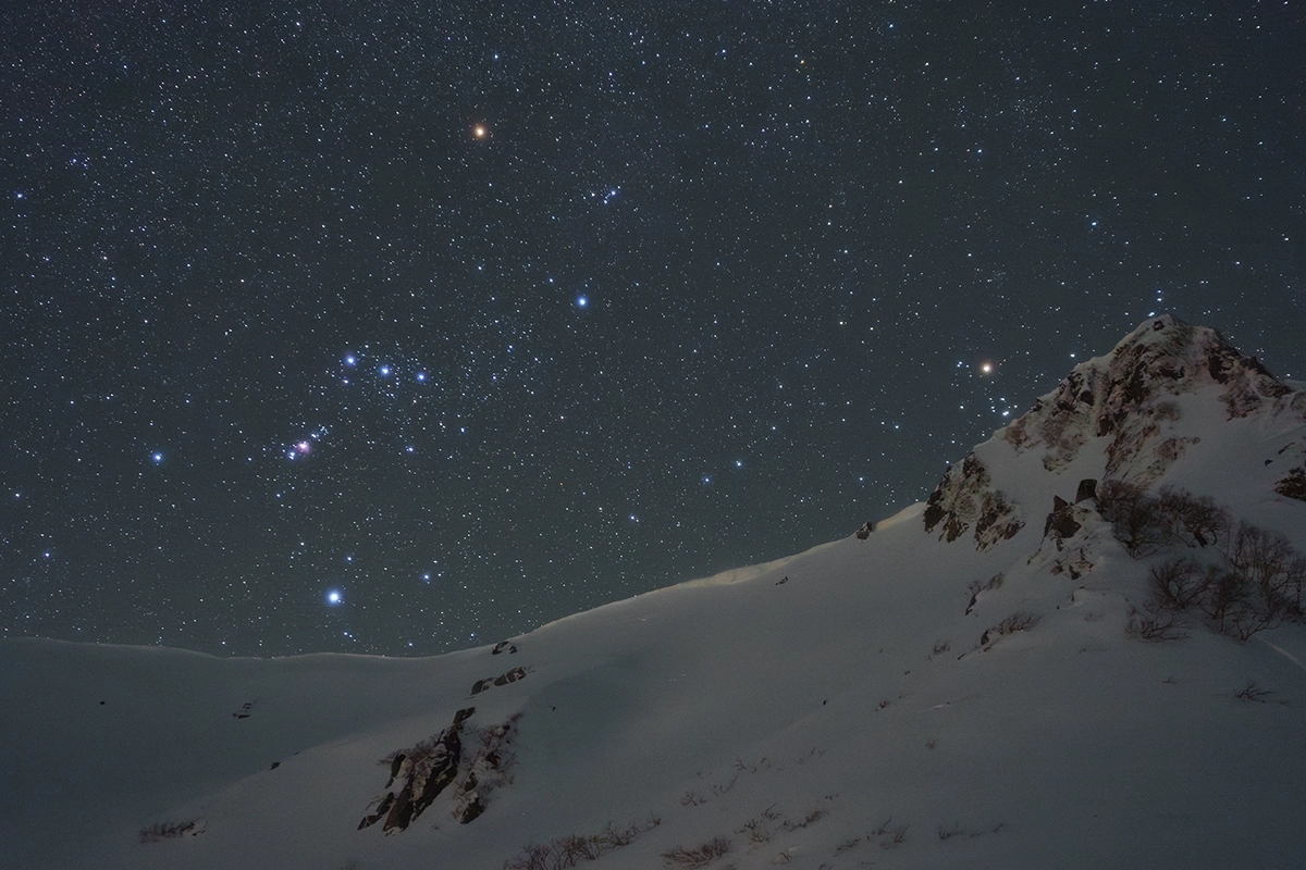 Cielo nocturno lleno de estrellas sobre una cresta montañosa nevada con la constelación de Orión visible, captada durante una sesión de astrofotografía invernal.