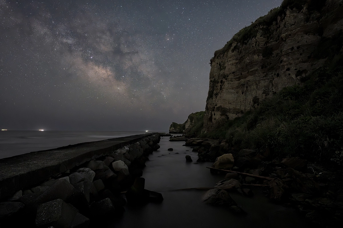 Galaxia de la Vía Láctea sobre un acantilado costero y un rompeolas, captada durante un paisaje nocturno de larga exposición que muestra las estrellas y la costa en silueta.