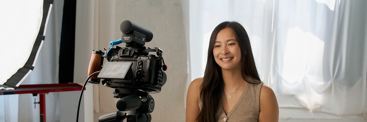 Mujer sentada delante de una cámara con un micrófono externo Sennheiser y un objetivo Tamron, grabando vídeo en un estudio con luz tenue.