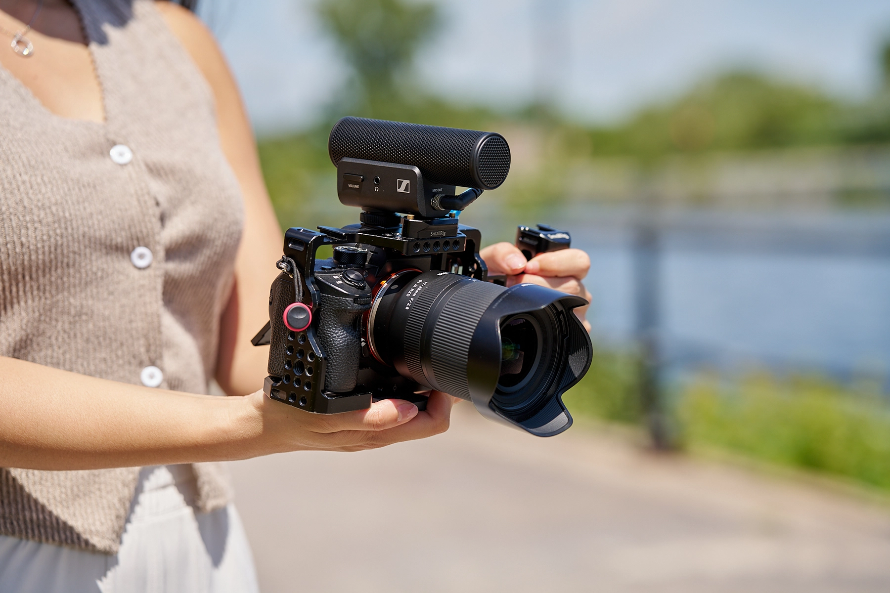 Persona sujetando una cámara sin espejo equipada con un objetivo Tamron y un micrófono externo Sennheiser, filmando en exteriores cerca de un paseo marítimo.