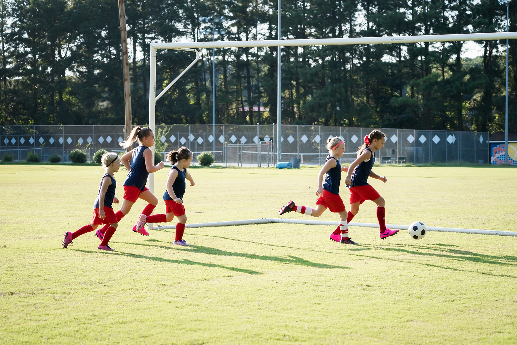 Un grupo de chicas jóvenes con uniformes de fútbol a juego persiguen el balón por un campo iluminado por el sol durante un partido, con expresiones concentradas y movimientos dinámicos capturados durante el juego.